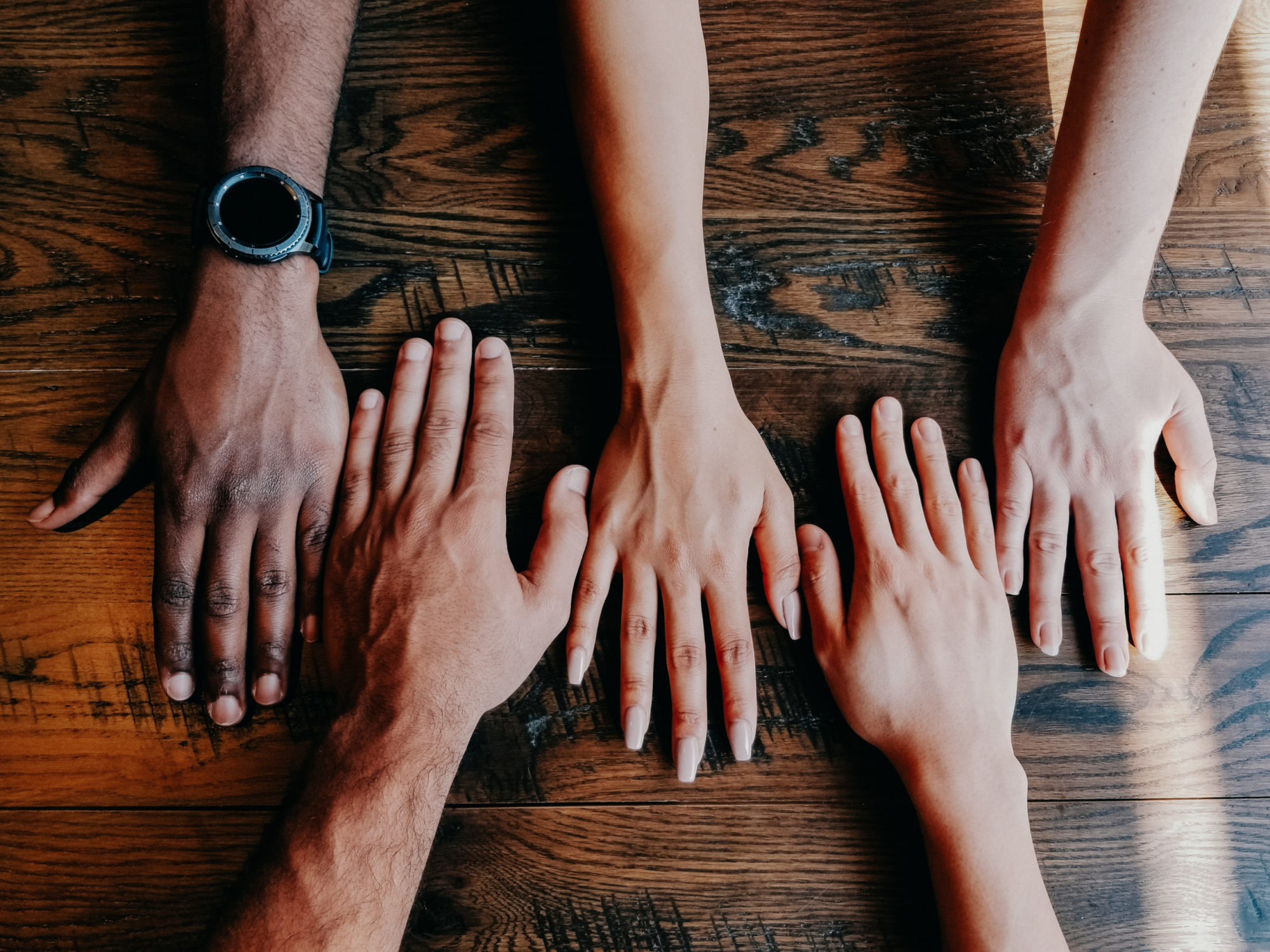 Hands of people from different races are laid next to each other in alternating directions, with the darkest skin color on the left in a gradient toward the lightest skin on the right.